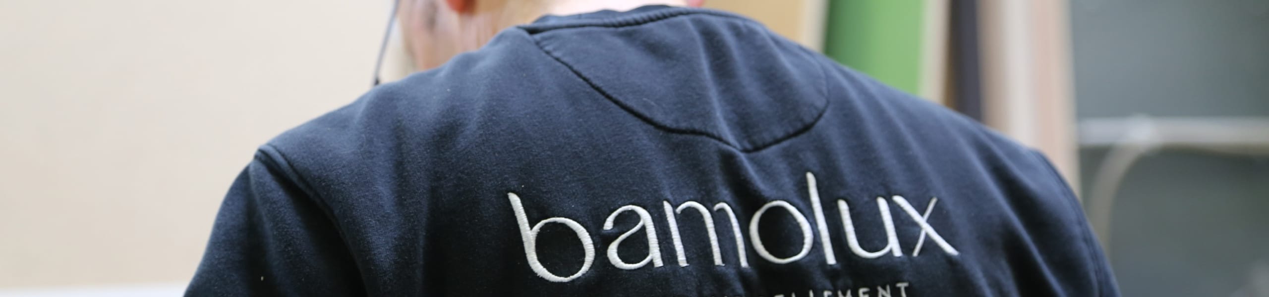 Menuisier vu de dos portant un t-shirt noir avec le logo ‘bamolux’, travaillant dans un atelier de menuiserie.